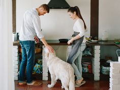 a man and woman standing next to a white dog on a tiled floor in front of a stove top oven