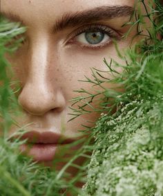 a woman with blue eyes is hiding her face behind some green plants and looking at the camera