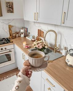 a woman is holding a cup of coffee in her hand while standing at the kitchen counter