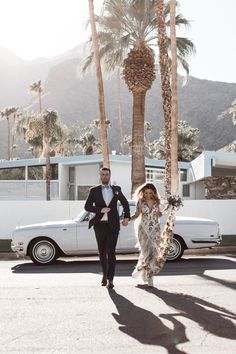 a bride and groom walking down the street in front of a vintage car with palm trees