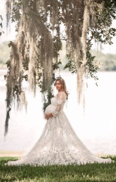 a pregnant woman in a wedding dress standing under a tree with moss hanging from it