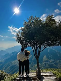 two people standing next to each other under a tree on top of a hill with mountains in the background