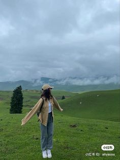 a woman standing on top of a lush green field under a cloudy sky with mountains in the background