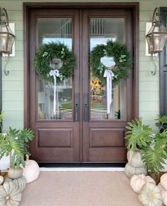 two wreaths on the front door of a house with pumpkins and plants in front