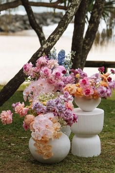 two white vases with flowers in them sitting on the grass near some trees and water