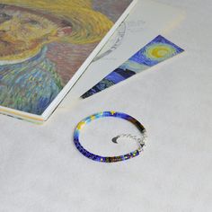 a beaded bracelet sits next to an open book on a white surface with a painting in the background