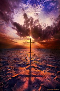 a cross in the middle of an empty field with clouds and sun setting behind it