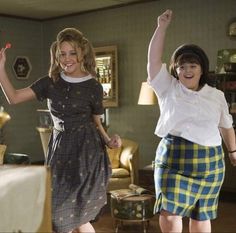 two women are dancing together in the living room, one is wearing a plaid skirt and the other wears a white shirt