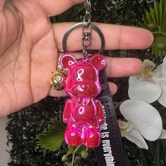 a pink bear keychain is being held by a person with flowers in the background