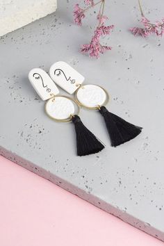 three white and black tasselled earrings on top of a pink table next to flowers