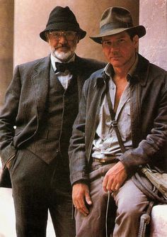 two men in suits and hats sitting next to each other on a stone bench outside