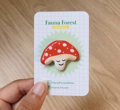 a hand holding a red and white mushroom brooch on top of a wooden table