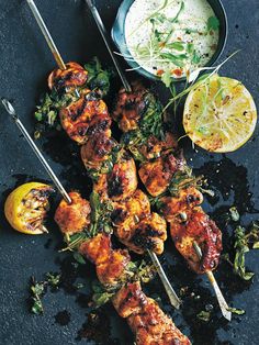 chicken skewers with herbs and lemons on a black surface next to a bowl of dip