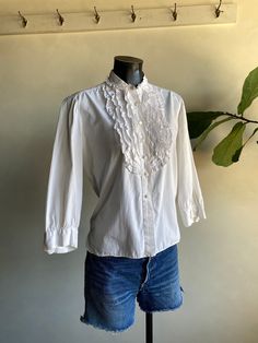 "Vintage 1970s Unbranded White Cotton Ruffle Blouse. White in color made from cotton fibers. Brand is unknown (tag has been cut). Long sleeve blouse with ruffled accents along front. Button down closure along front and cuffs. Overall condition of garment is good with only minor flaws that include; blouse is missing one of the buttons on the right cuff as shown in photos. No rips, tears, or staining. Please see all photos for details. Refer to measurements below to ensure a proper fit (modeled on Vintage Shirt For Spring Daywear, Vintage Shirt For Daywear In Spring, Vintage White Cotton Blouse For Spring, Fitted Cotton Blouse For Vintage Fashion, Classic Cotton Tops For Vintage Fashion, Vintage Cotton Tops For Daywear, Vintage Ruffled Tops For Spring, Victorian Cotton Tops For Spring, Vintage White Cotton Top For Spring