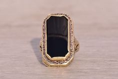 This stunning piece, known as The Art Deco Elegance, features a captivating elongated emerald cut onyx tablet at its center. The rectangular cut onyx is elegantly bezel set in 14 karat yellow gold, designed with intricate filigree detailing that adds a touch of vintage sophistication which continues down the shank. The ring is crafted in 14 karat yellow gold and is currently a finger size 2.75. Art Deco Onyx Ring, Elegant Formal Onyx Signet Ring, Elegant Intaglio Signet Ring, Elegant Rectangular Jewelry With Polished Edges, Elegant Black Enamel Rectangular Jewelry, Elegant Onyx Signet Ring With Gemstone, Elegant Rectangular Black Enamel Jewelry, Elegant Black Rectangular Signet Ring, Elegant Formal Signet Ring With Intaglio