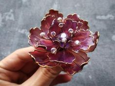 a hand holding a flower shaped brooch with pearls on it's center, in front of a gray background