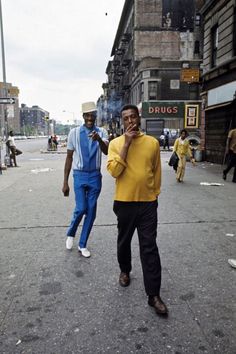 two men are walking down the street talking on their cell phones and one is wearing a yellow shirt