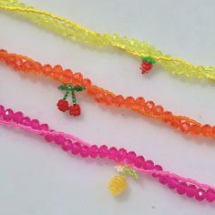 four different colored bracelets on a white surface with beads and fruit charms hanging from them