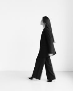 a woman walking across a white floor with her hair blowing in the wind and wearing a black suit