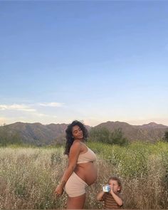 a pregnant woman standing next to a little boy in a field with mountains behind her