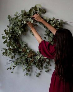 a woman in a red dress is hanging a wreath on the wall with greenery