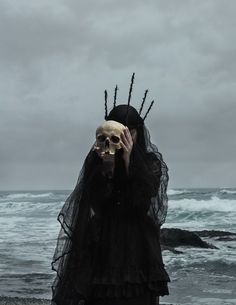 a woman in a black dress holding a human skull on her head while standing next to the ocean