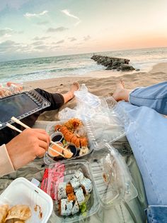 two people are sitting on the beach eating sushi and chopsticks with their feet in the sand