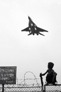 a boy sitting on a fence looking at an airplane in the sky above him, with barbed wire behind him