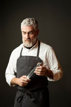 an older man with grey hair and beard wearing black apron holding a knife in his hands