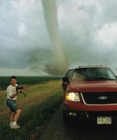 a man standing next to a red truck on a road with a large tornado in the background