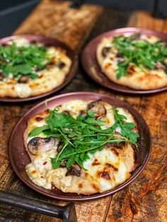 three small pizzas with greens on them are sitting on a wooden table next to a knife and fork