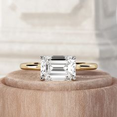 an emerald - cut diamond ring sits on top of a wooden stand in front of a white marble wall