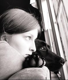 a woman holding a dog's paw up to her face while looking out the window