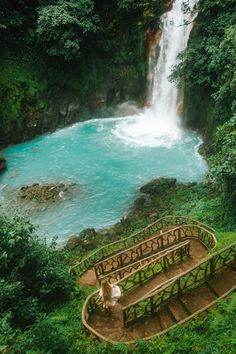 stairs lead down to a waterfall in the middle of green trees and blue water below