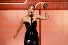 a woman in a black dress is holding a bottle and smiling at the camera while standing on stage