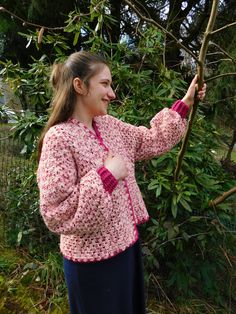 a woman holding onto a tree branch while wearing a pink and white knitted jacket