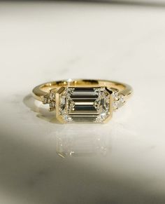 an emerald cut diamond ring with three smaller diamonds on the band, set in yellow gold