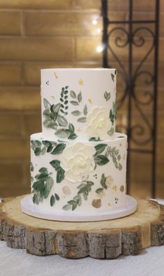 a wedding cake decorated with white flowers and green leaves on a wooden slice in front of a brick wall