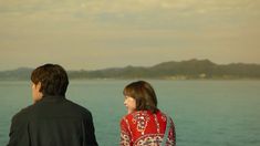 a man and woman standing next to each other looking at the ocean with mountains in the background