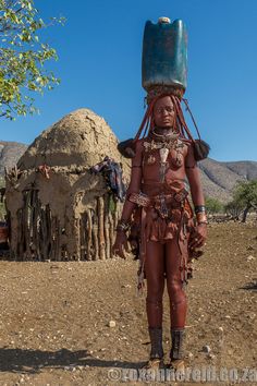 an african woman in traditional garb and headdress stands outside her hut, holding a bucket on her head