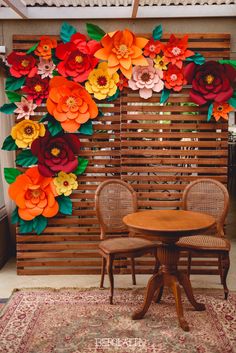 an arrangement of paper flowers on the wall behind two chairs and a round table in front of a wooden slatted screen