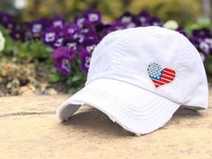 a white baseball cap with an american flag heart patch on it sitting in front of purple flowers