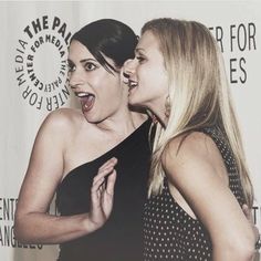 two beautiful women standing next to each other in front of a white wall with words on it