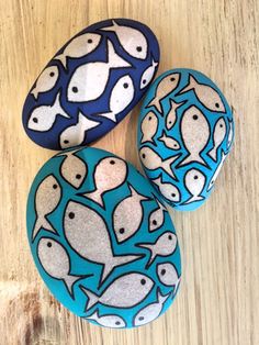 three painted rocks sitting on top of a wooden table covered in blue and white fish