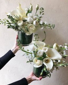 two hands holding vases with white flowers in them and greenery on the side