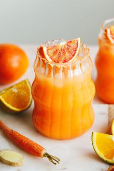 an orange drink in a glass pitcher surrounded by citrus fruits and vegtables