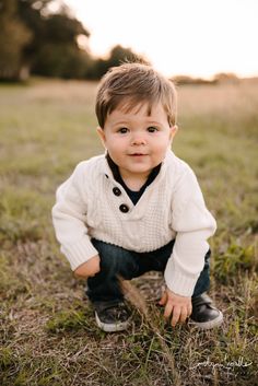 a little boy sitting in the grass with his hands on his knees and looking at the camera