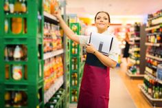 a woman in an apron is holding up a piece of paper while standing next to a grocery aisle