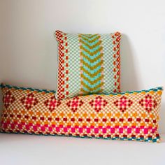 two colorful pillows sitting next to each other on top of a white table with a wall in the background