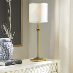 a table lamp sitting on top of a white dresser next to a book and vase